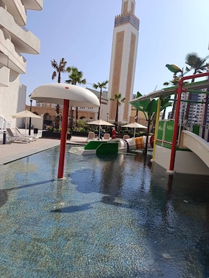 Een zwembadgedeelte met helder water, ligbedden, parasols en een hoog gebouw op de achtergrond.
