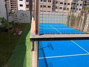 Een tennisbaan met een net, omgeven door een metalen hek, en een bank in een aangelegde tuin.