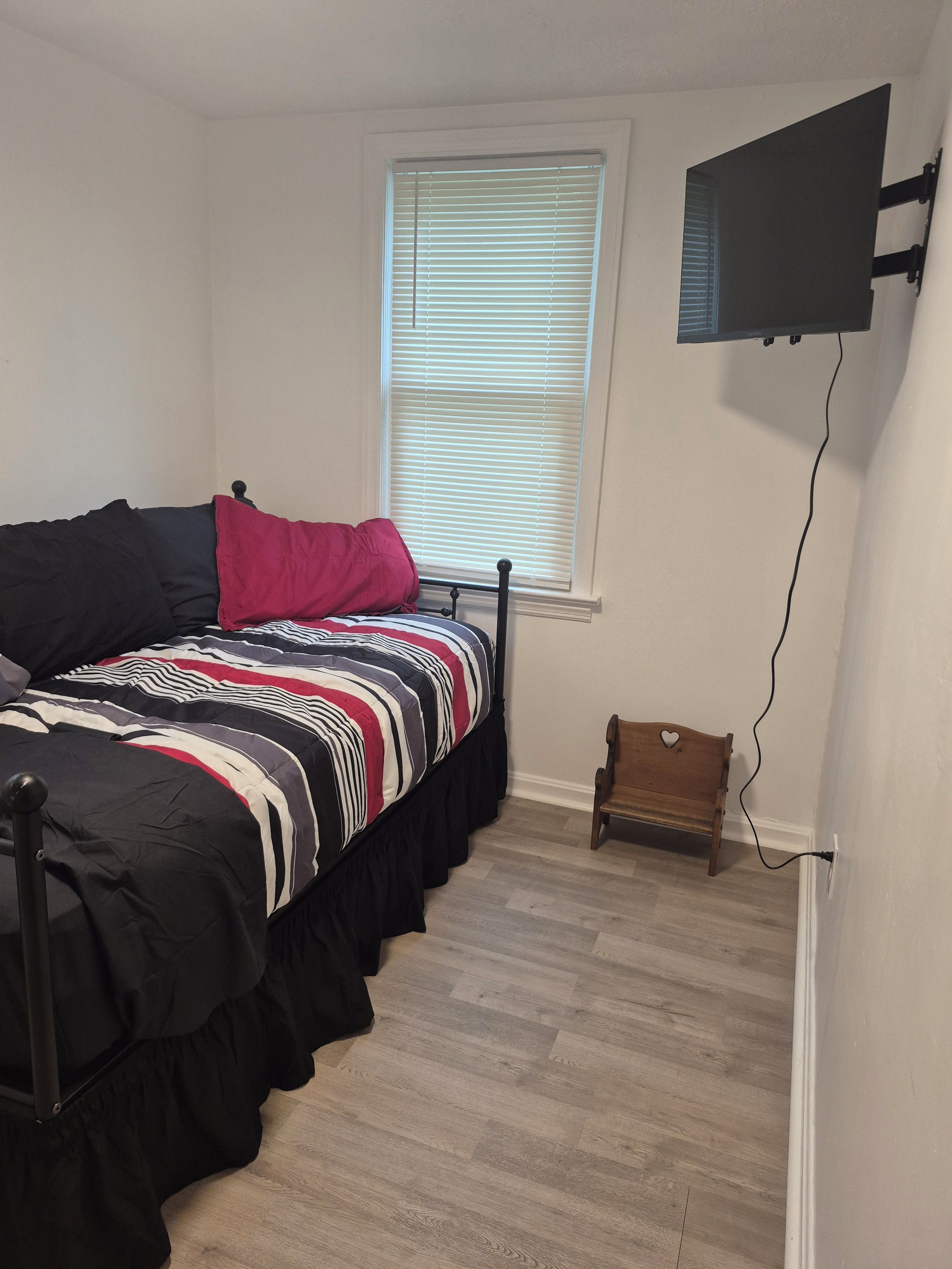A bedroom with a bed, a television mounted on the wall, a window with blinds, and a small wooden chair.