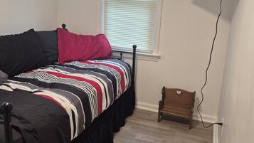 A bedroom with a bed, a television mounted on the wall, a window with blinds, and a small wooden chair.