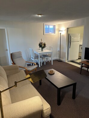 A living room with a sofa, a coffee table, and a dining table with chairs. There is a refrigerator and a TV in the background. - Central Oakville/Kerr Village:steps to shops, eateries, cinema, transit, & GO  (Oakville)