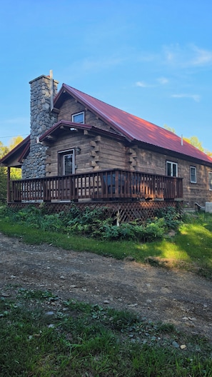 A log cabin with a stone chimney, wooden deck, and a metal roof. - Cozy 3 bedroom with loft cabin in serene Pittsburg with AC & woodstove. (Pittsburg)