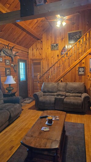 A cozy cabin interior with a wooden staircase, a sofa, a coffee table, and a ceiling fan. - Cozy 3 bedroom with loft cabin in serene Pittsburg with AC & woodstove. (Pittsburg)