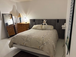 Un dormitorio con una cama, un aparador, un espejo, una lámpara y un peluche.