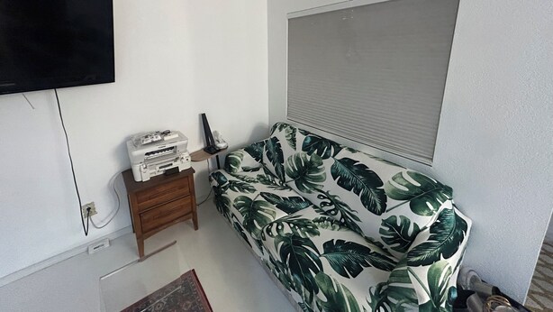 Een kamer met een bed, een aan de muur bevestigde televisie, een klein houten nachtkastje en een raam met jaloezieën.