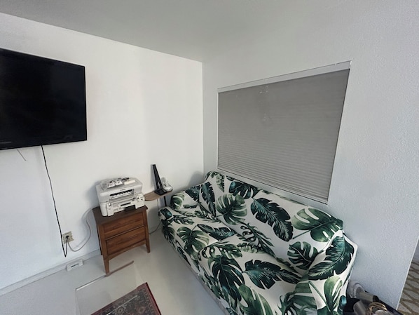 Una habitación con una cama, un televisor montado en la pared, una mesita de noche de madera y una ventana con persianas.