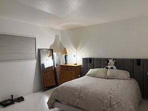 Un dormitorio con una cama, un aparador, un espejo, una lámpara y un peluche.