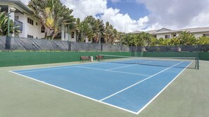 Een tennisbaan buiten met net, omgeven door een groene hekken en palmbomen.