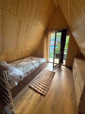 A single-bed room with a wooden sloped ceiling, a small bedside table, a window with curtains, and a view of a building outside. - Getamej Eco Villa & Pool (Getamej)