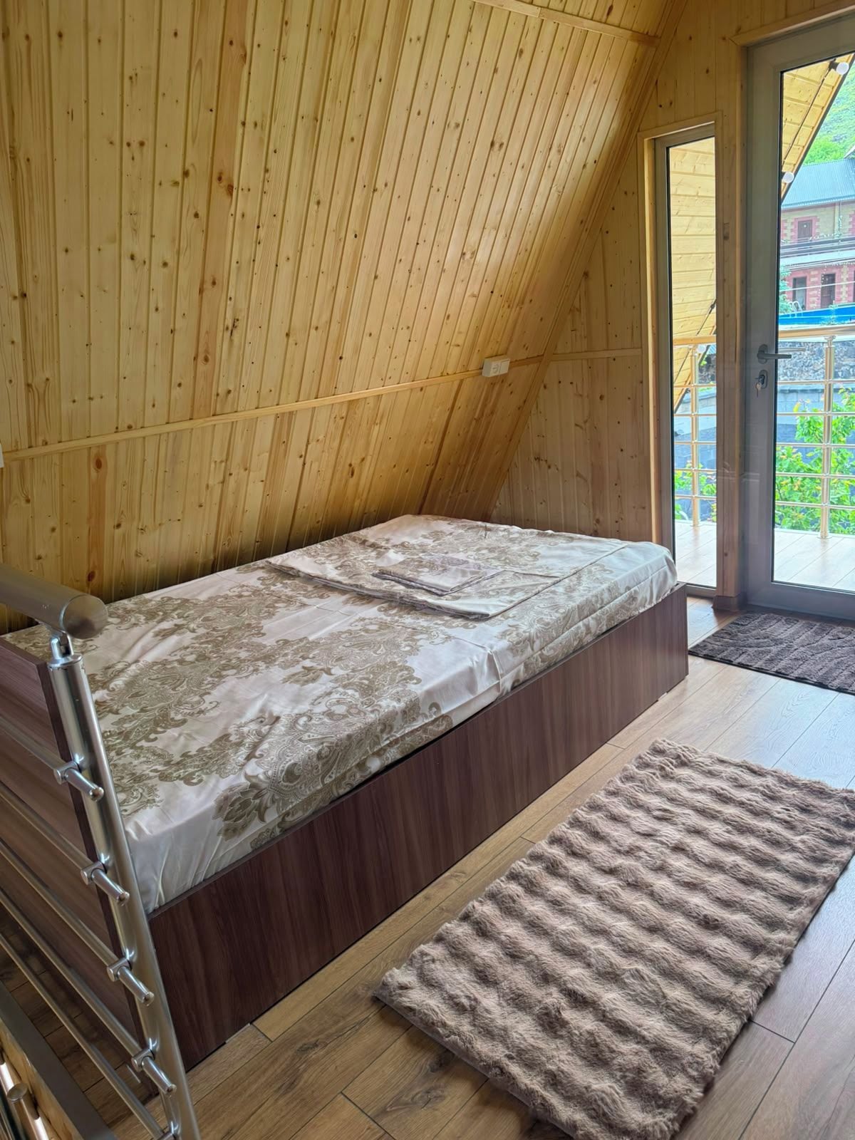 A small, wooden room with a single bed, a wooden staircase, and a view through a glass door.