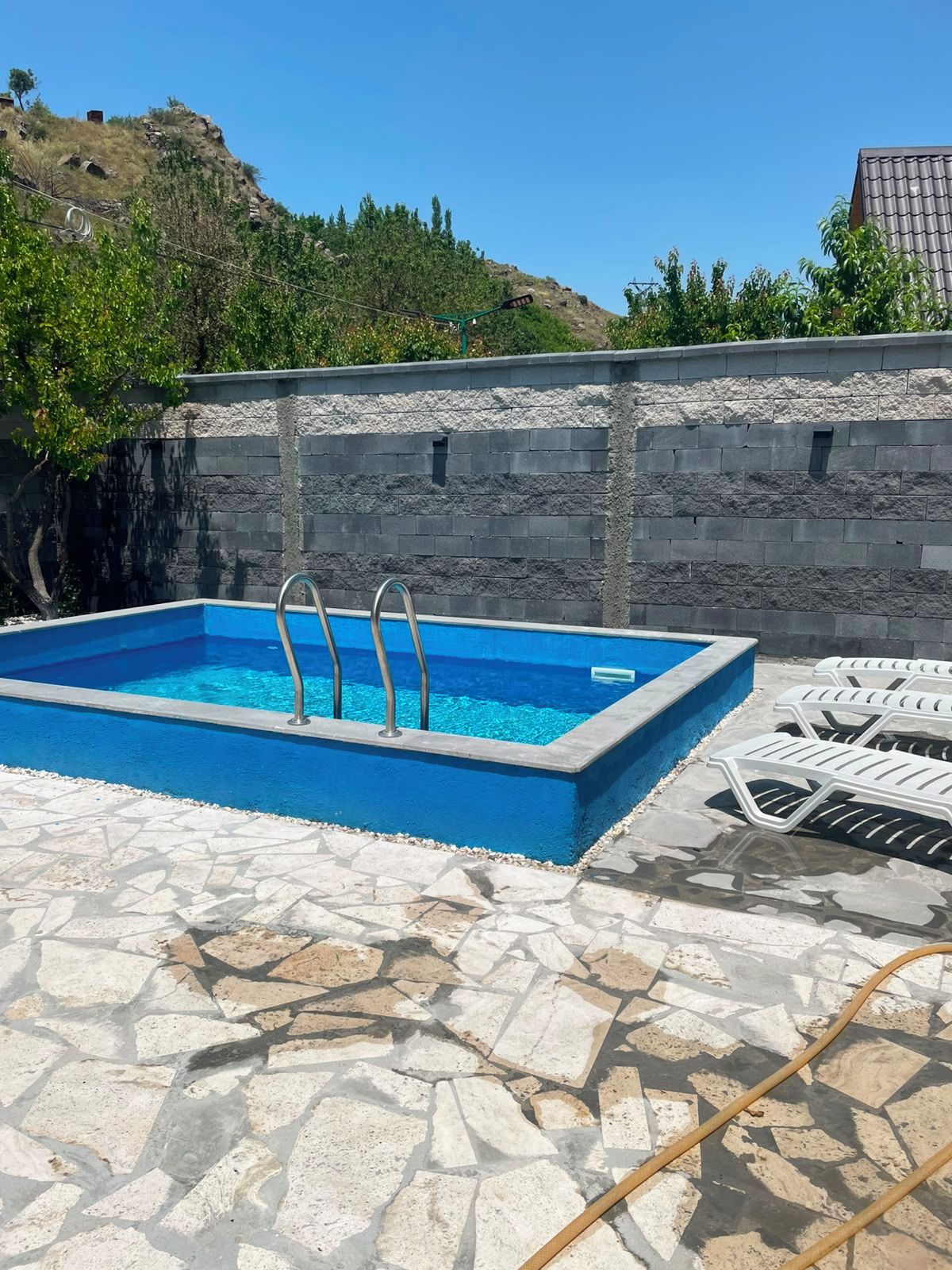 Una piscina rectangular al aire libre con dos escaleras, rodeada de baldosas de piedra y un muro de piedra.