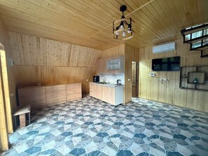 A room with a sloped ceiling, a built-in kitchen, a stone-patterned floor, and a television mounted on the wall.