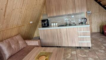 Cozinha compacta com armários de madeira e bancada, uma mesa de jantar com um objeto decorativo dourado, e um sofá em um quarto com paredes de madeira inclinadas.