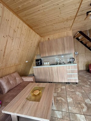 A compact kitchen with wooden cabinets and a countertop, a dining table with a gold decorative item, and a sofa in a room with sloped wooden walls. - Getamej Eco Villa & Pool (Getamej)
