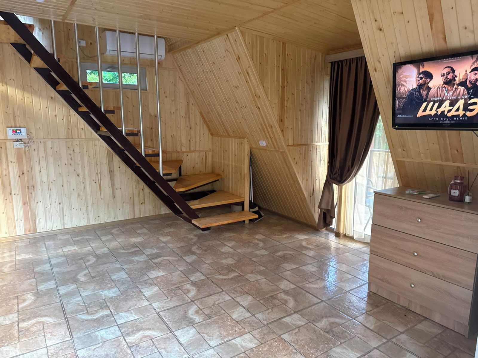 Una habitación con una escalera de madera, un televisor en la pared y piso de baldosas.