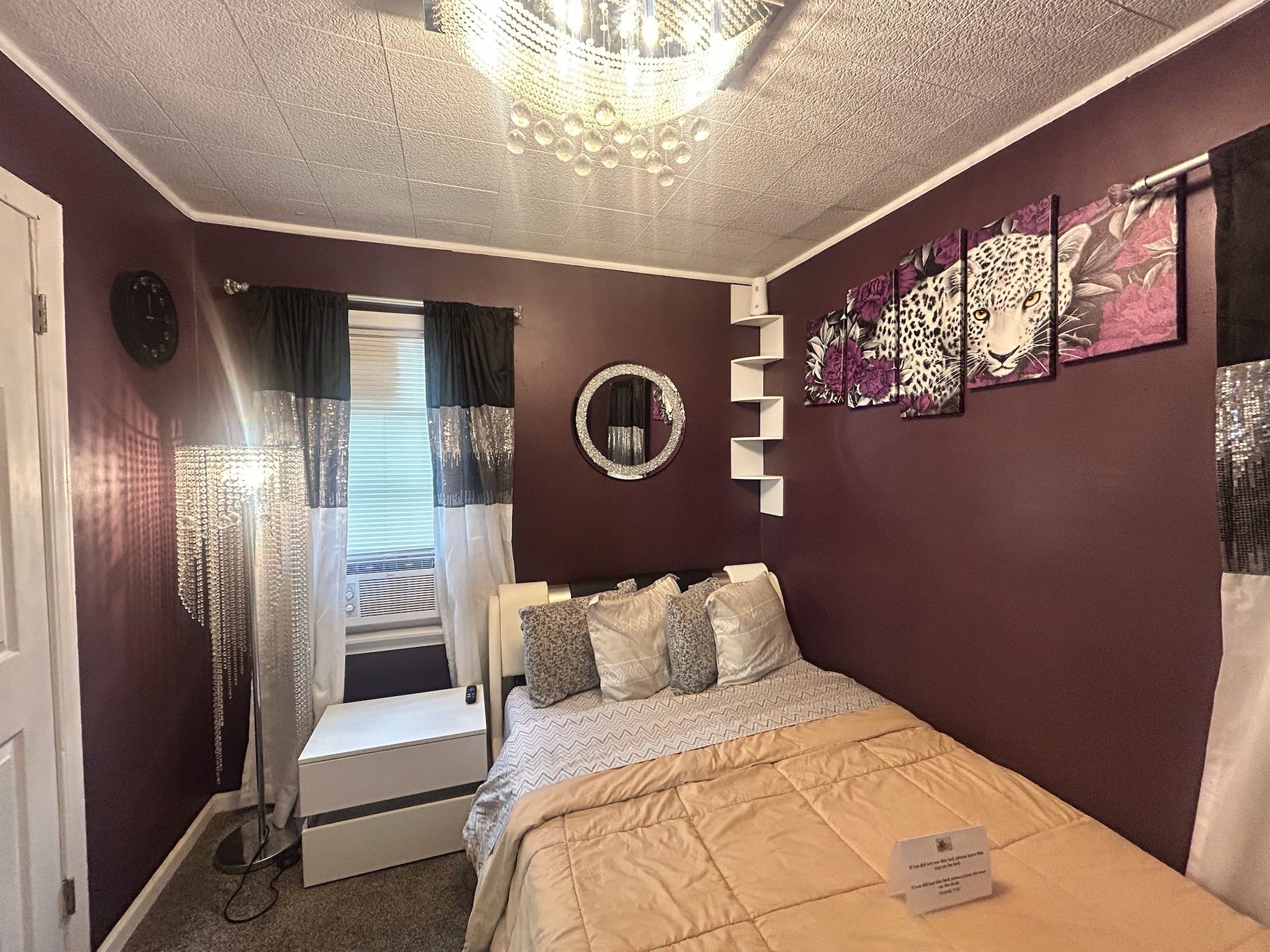 Een slaapkamer met een bed, nachtkastje, spiegel en raam met gordijnen.