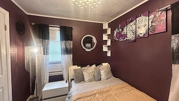 Een slaapkamer met een bed, nachtkastje, spiegel en raam met gordijnen.