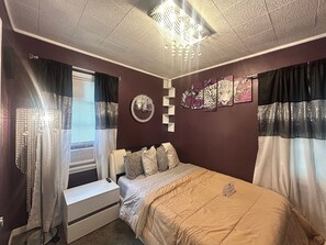 Quarto com cama, mesa de cabeceira, luminária, janela com cortinas e decoração na parede.