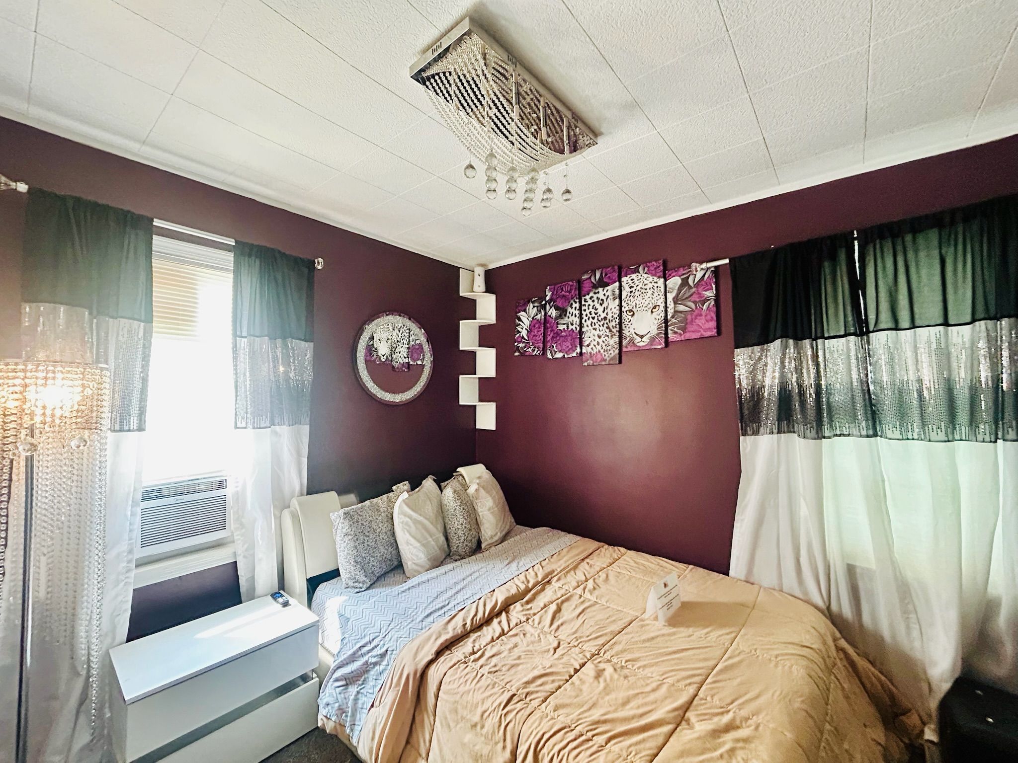 Un dormitorio con cama, ventana con cortinas, aire acondicionado de pared y decoración en la pared.