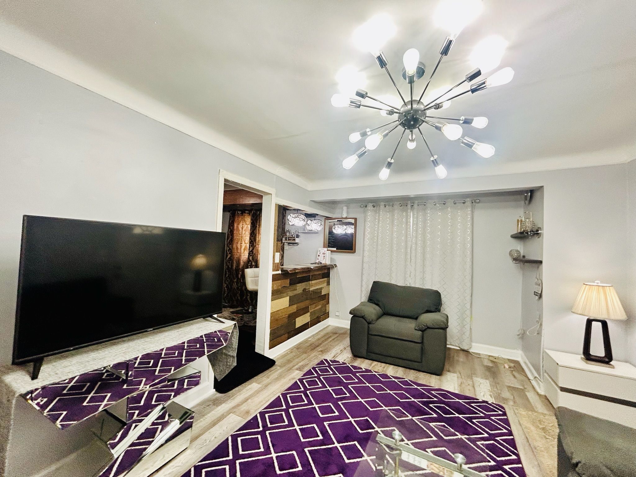 Una sala de estar con una televisión de pantalla plana, un sofá, un tapete estampado, un moderno accesorio de luz de techo y una mesita con una lámpara.