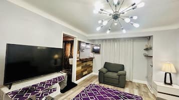 Una sala de estar con una televisión de pantalla plana, un sofá, un tapete estampado, un moderno accesorio de luz de techo y una mesita con una lámpara.