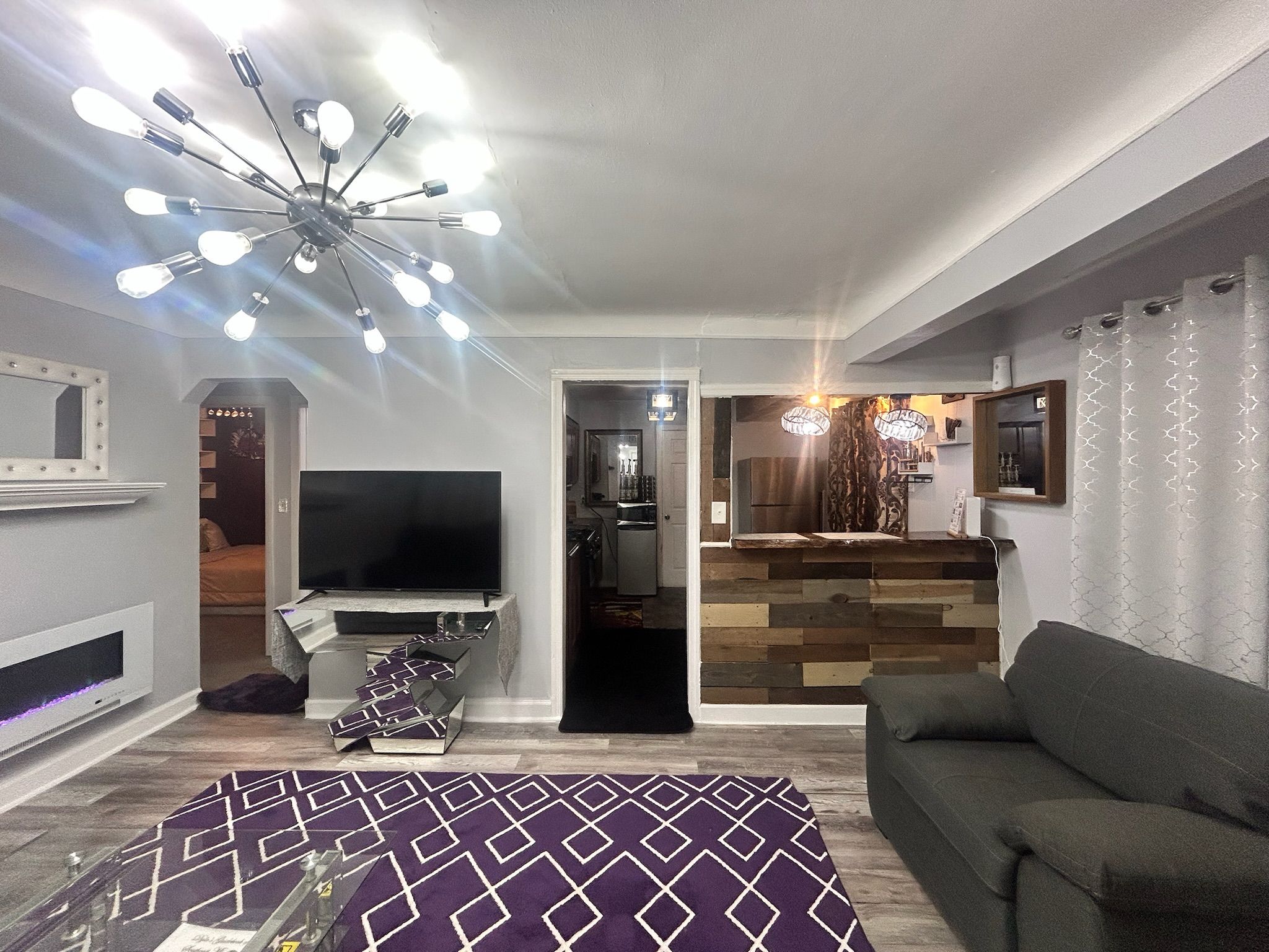 Una sala de estar con televisor, sofá, alfombra estampada y una lámpara moderna en el techo.