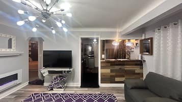 Una sala de estar con televisor, sofá, alfombra estampada y una lámpara moderna en el techo.