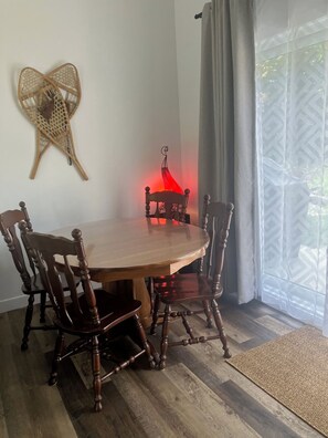 A dining area with a round wooden table and four chairs, a woven wall hanging, and a view through a window with curtains. - Chalet with 2 bedrooms, 5 minutes from Mount Adstock. Skiing, golf, hiking (Adstock)