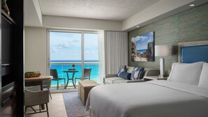 Quarto de hotel com uma cama grande, varanda com vista para o mar, área de refeições com mesa e cadeiras, e uma televisão.