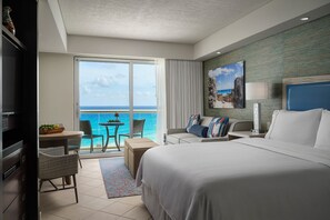 Una camera d'albergo con un letto grande, un balcone vista oceano, un'area pranzo con tavolo e sedie e una televisione.