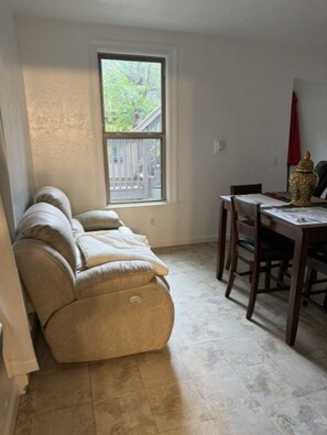 Una habitación con un sofá color crema, una mesa de comedor con sillas y una ventana con vista a árboles.