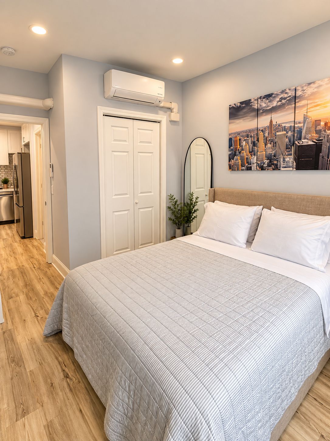 A bedroom with a bed, a cityscape painting, a mirror, and an air conditioning unit.