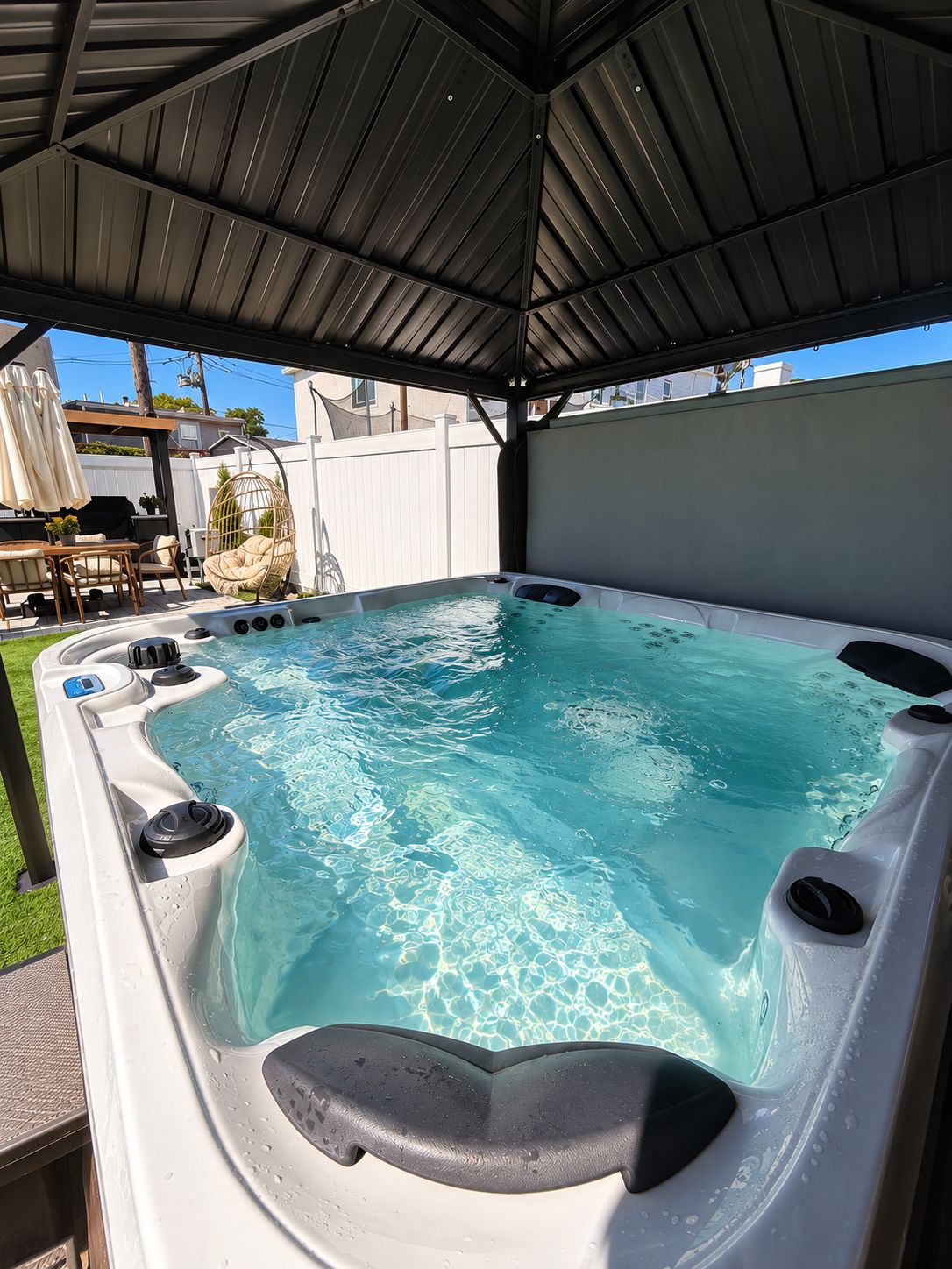 A hot tub with a metal roof structure, surrounded by a white fence, and a patio area with outdoor furniture.