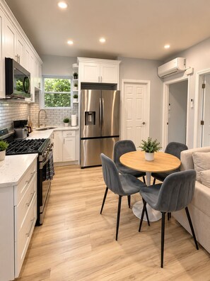 A modern kitchen with white cabinets, stainless steel appliances, a round wooden table with chairs, and a beige sofa in the adjacent living area.