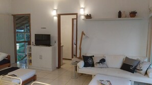 A living room with a sofa, a television on a cabinet, a bedside table, and a window with a view of greenery. - House with ocean view in Ilhabela (Ilhabela)