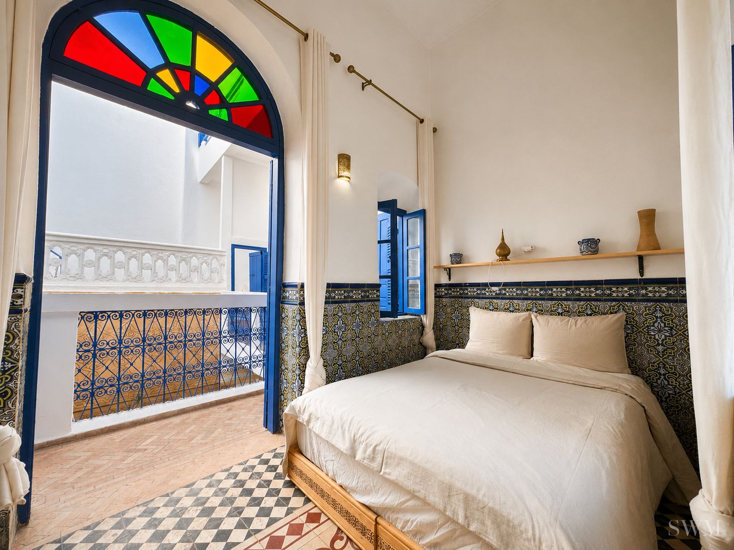 A bedroom with a bed, a window with blue shutters, a tiled floor, and a balcony with a railing.