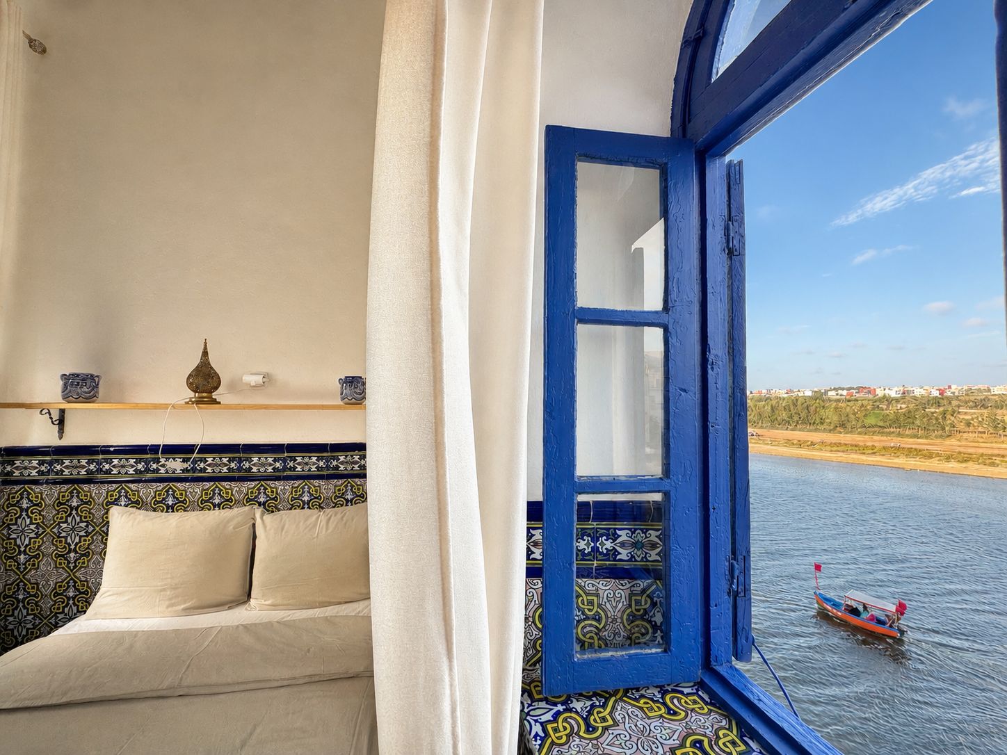 A room with a bed, a window with blue trim, and a view of a river with a boat.