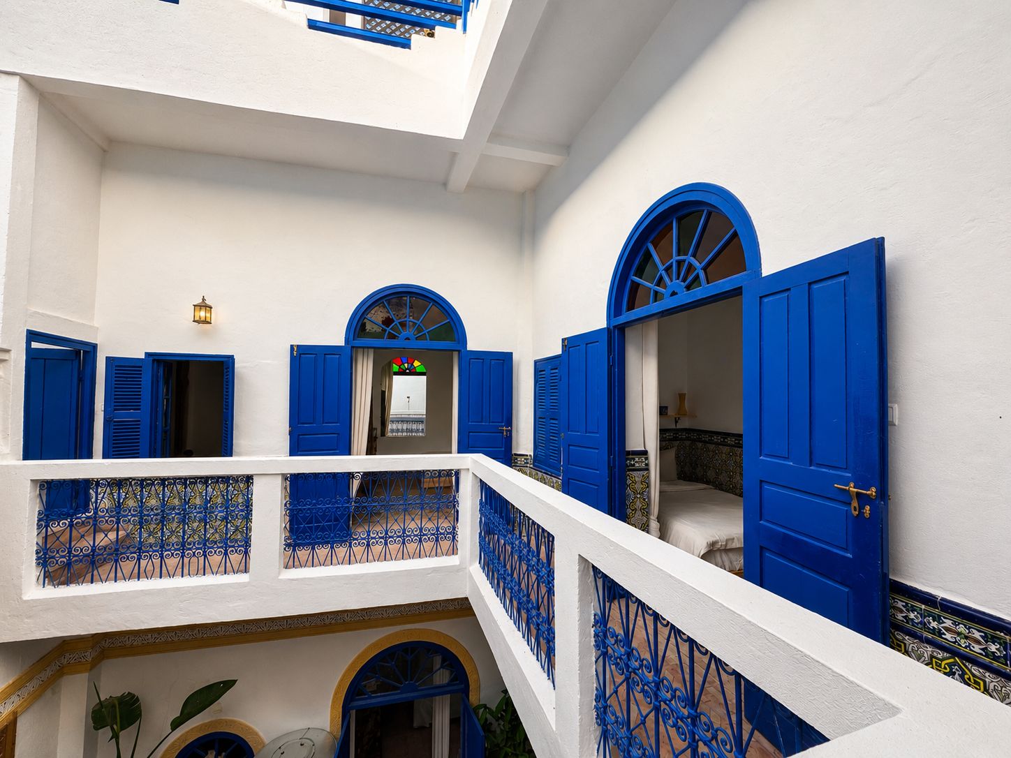 Un edificio blanco con puertas y contraventanas azules, un balcón con barandilla azul y un suelo de baldosas.