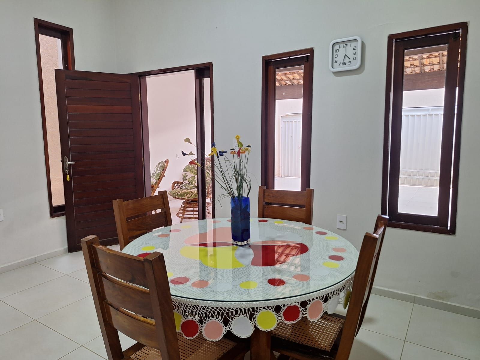 Un comedor con una mesa redonda, sillas, un florero con flores y un reloj en la pared.