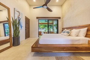 A bedroom with a large bed, a ceiling fan, a potted plant, and a view of the outdoors through a sliding door.