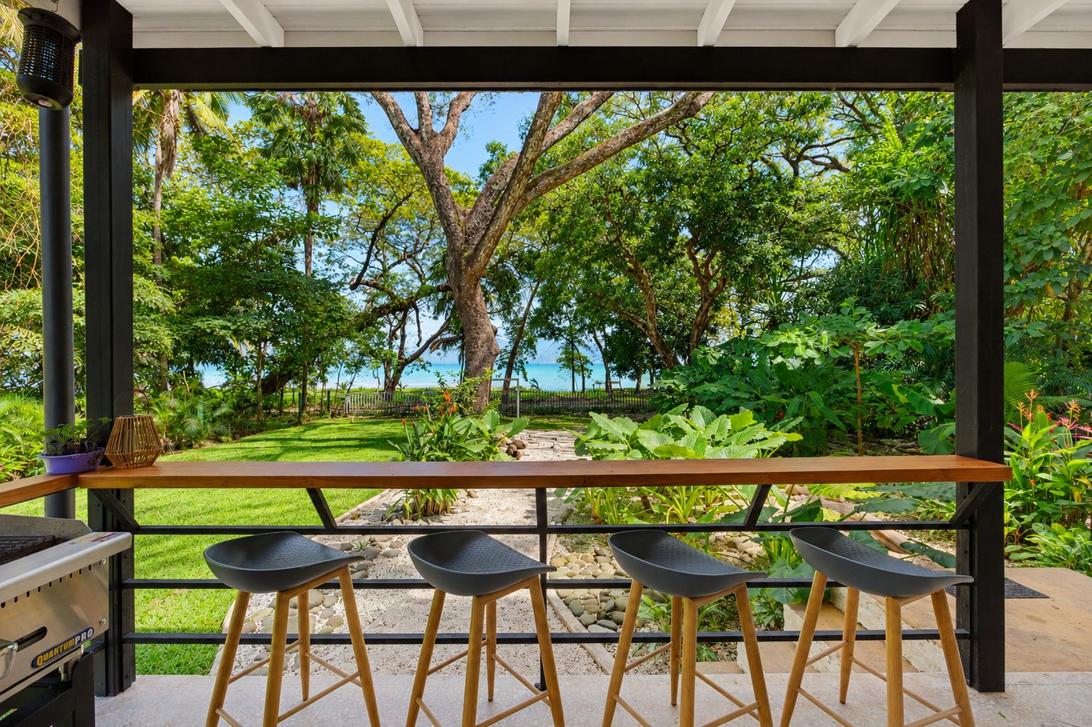 An outdoor seating area with a grill and stools, overlooking a lush garden and a view of the sea.