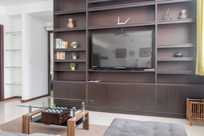 Uma sala de estar moderna com uma TV de tela plana presa a uma parede de madeira escura, uma mesa de centro de vidro com um jogo de xadrez e um sofá cinza.