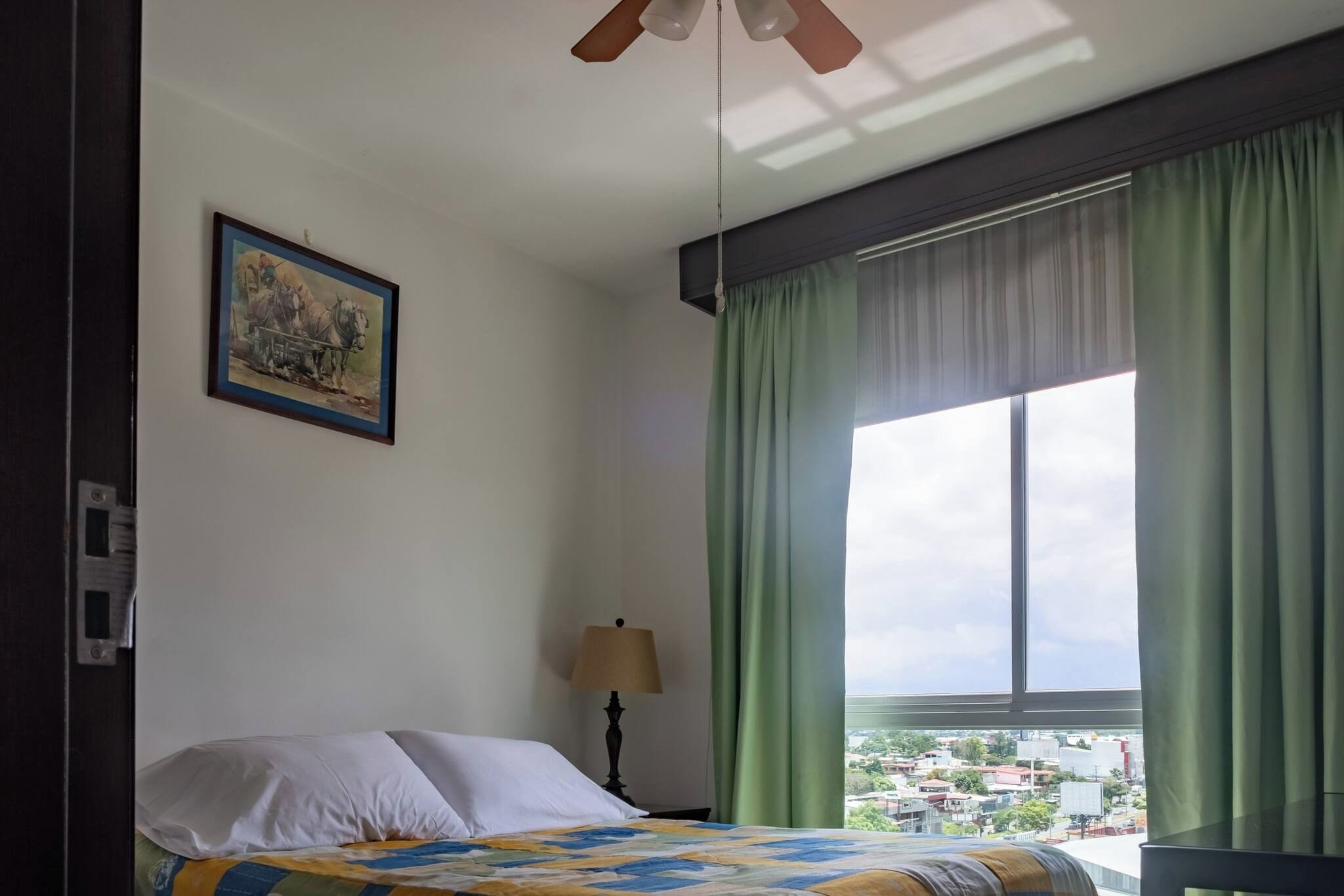 A hotel room with a bed, a painting, a window with curtains, and a view of the city.