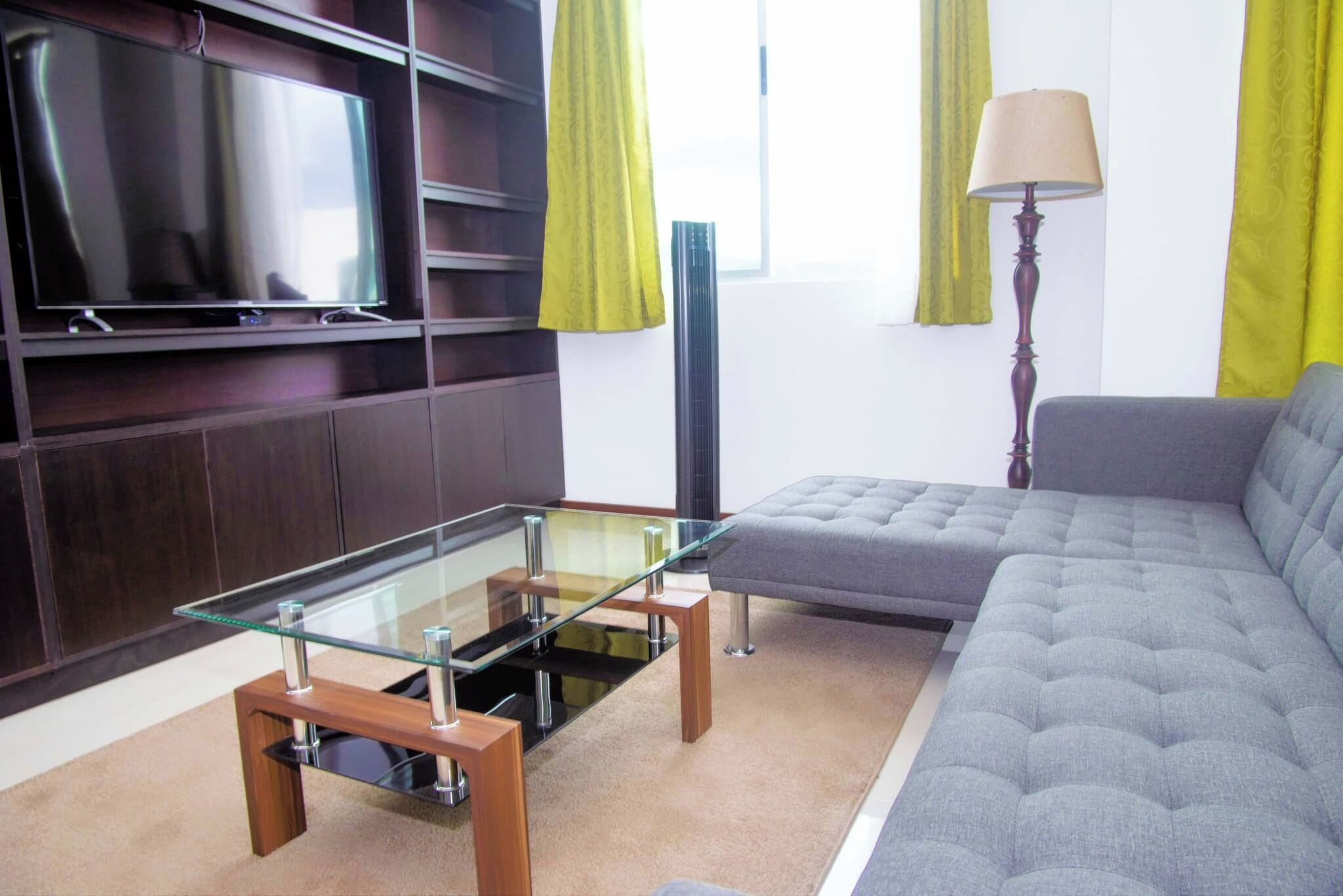 Un salon moderne avec une table basse en verre, un canapé gris, un téléviseur à écran plat et un grand lampadaire.