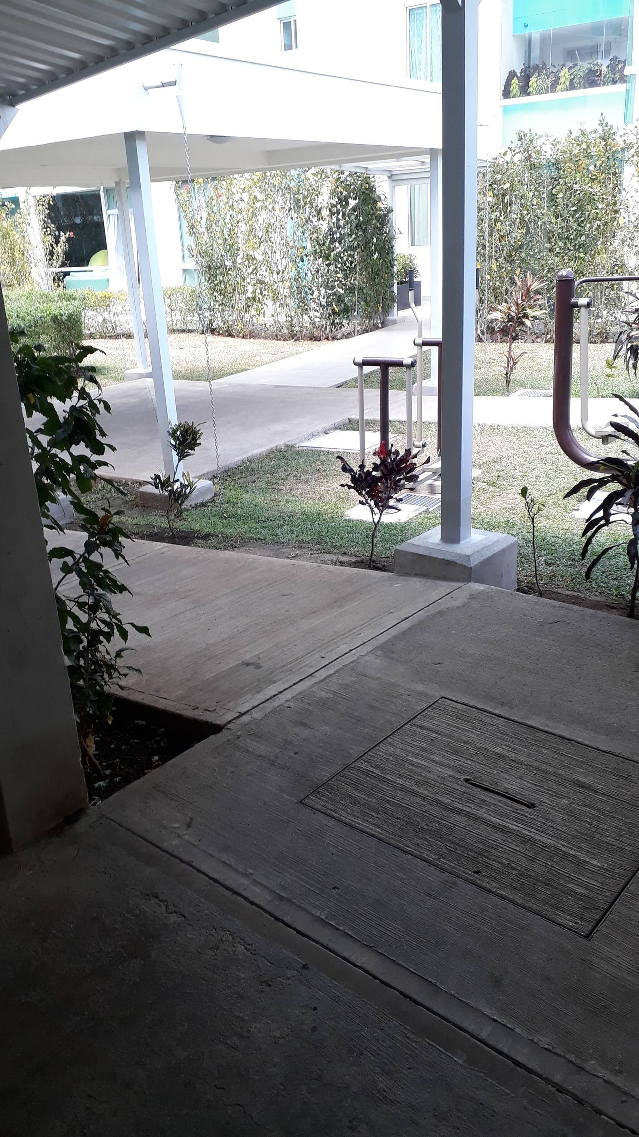 A covered outdoor area with a concrete floor, a metal bench, and a small garden with plants.