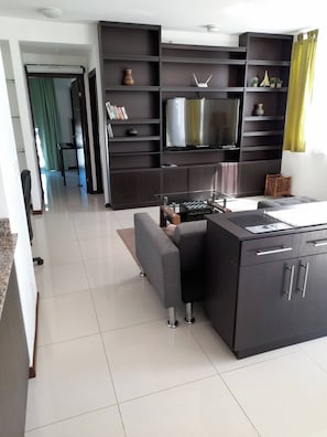 A modern kitchen with a built-in stove, a dining area with a sofa, and a flat-screen TV mounted on a dark wooden wall.