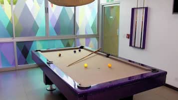 Une table de billard avec des billes et une queue, dans une pièce dont les fenêtres présentent des motifs géométriques aux couleurs vives.