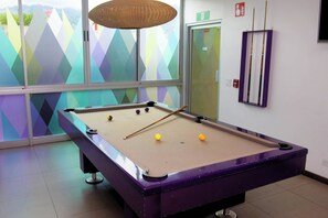 Une table de billard avec des billes et une queue, dans une pièce dont les fenêtres présentent des motifs géométriques aux couleurs vives.