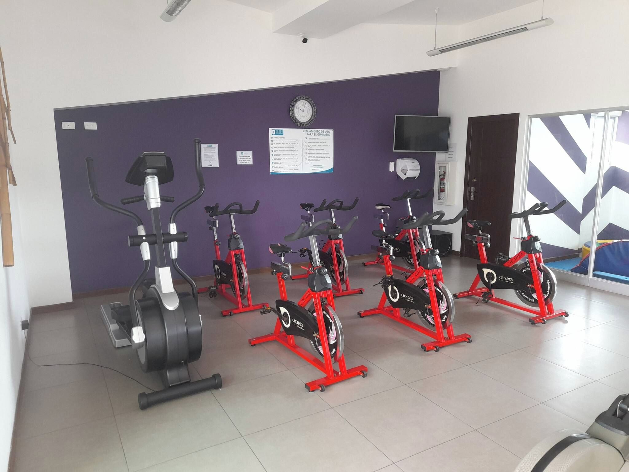 Ein Fitnessraum mit in Reihen angeordneten Ergometer, einer Wanduhr und einem Fernseher über einer Tür.