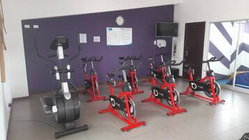 Ein Fitnessraum mit in Reihen angeordneten Ergometer, einer Wanduhr und einem Fernseher über einer Tür.